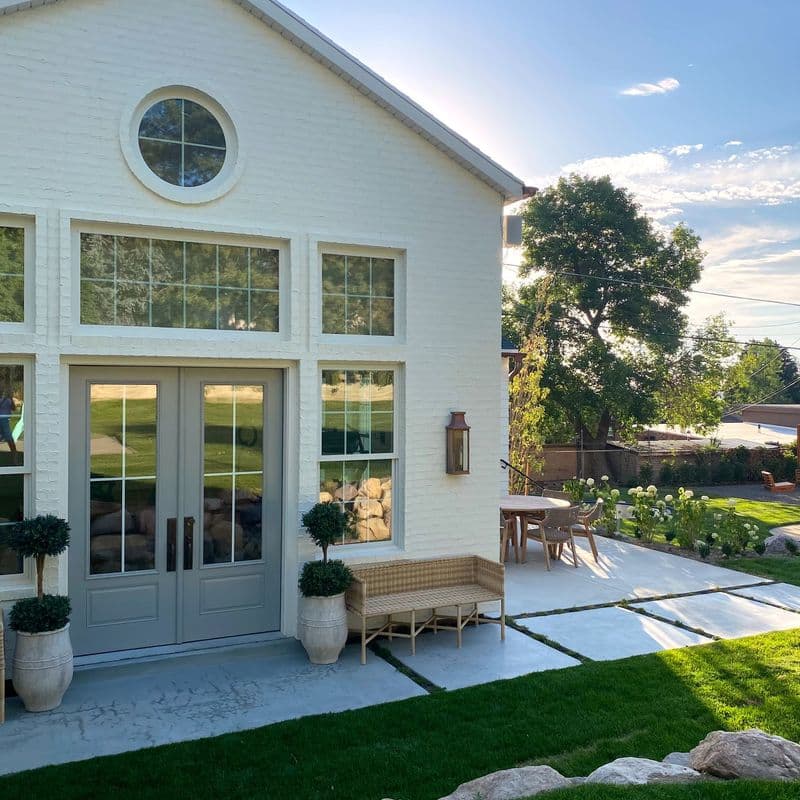 White brick house with large windows and a circular window. French doors open to a patio with potted plants, a wicker bench, and outdoor furniture. Landscaped lawn with concrete pavers and decorative rocks. Trees and blue sky visible in the background.