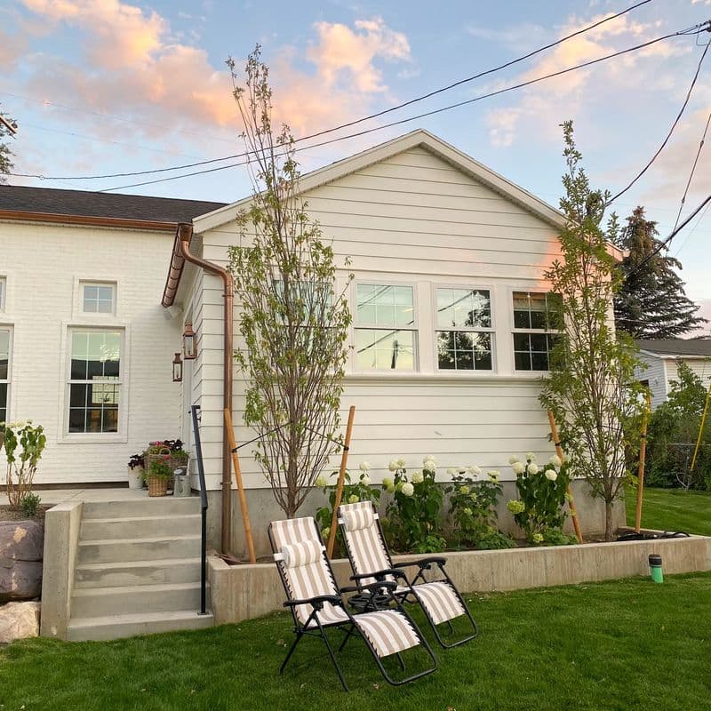 White suburban house with a newly landscaped front yard featuring young trees, a raised garden bed, and two striped lounge chairs on a green lawn. The scene is bathed in warm sunset light with power lines visible overhead.