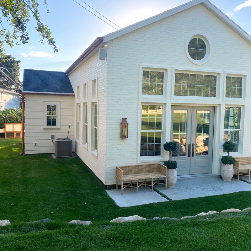 White brick house with large windows and a circular window. French doors open to a patio with wicker bench and potted plants. Landscaped lawn with rocks bordering the patio. A smaller connected structure is visible to the left with an air conditioning unit.