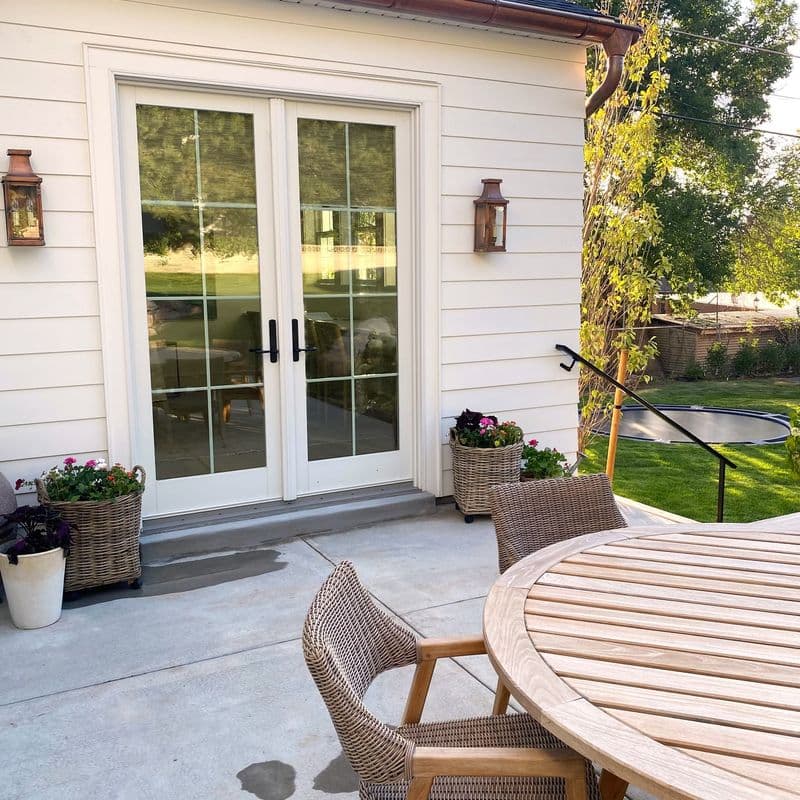 White house exterior with French doors, copper gutters, and lantern sconces. Concrete patio features wicker chairs, a wooden table, and potted flowers. A lawn and trees are visible in the background.