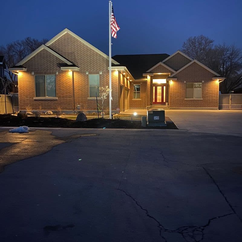 A suburban home at night, with the exterior lights illuminating the brick facade and American flag on a pole in the front yard. The driveway and street are also visible in the foreground.