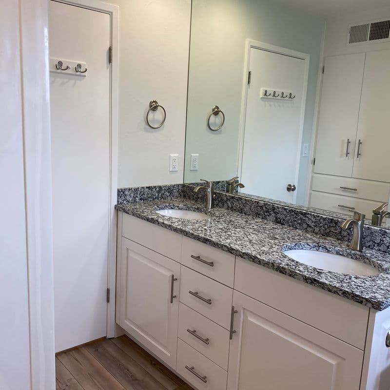 A modern bathroom with a large mirror, white cabinets, a gray granite countertop with double sinks, and chrome fixtures.