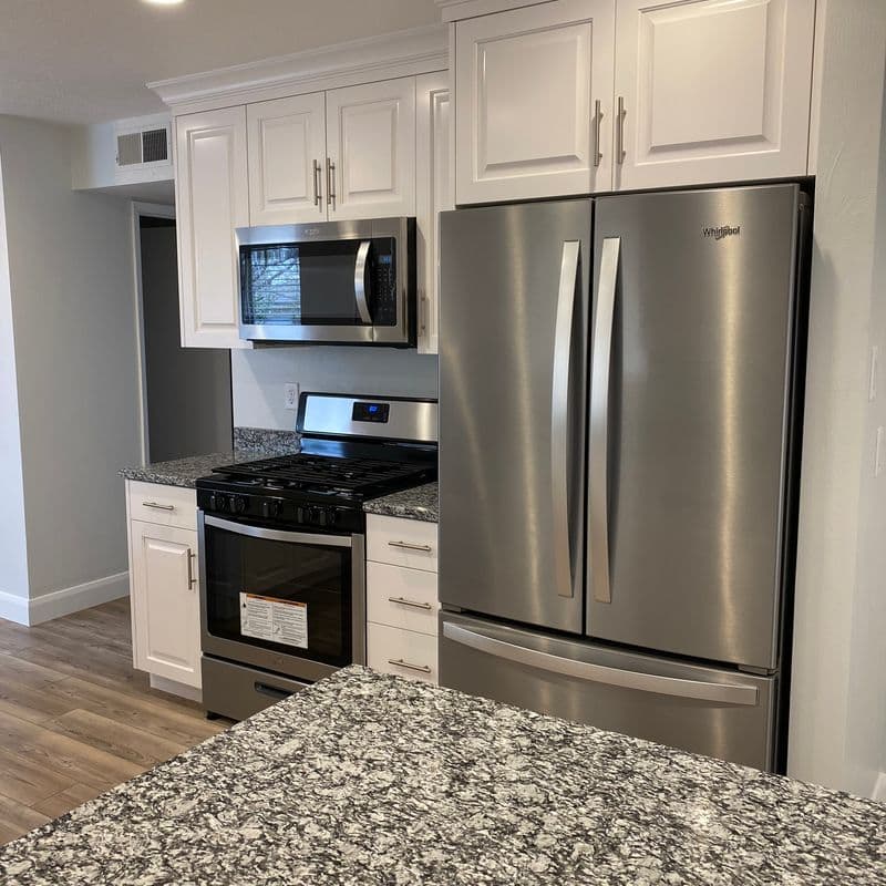 Stainless steel appliances including a microwave and refrigerator built into white cabinets above a gray granite countertop in a modern kitchen.