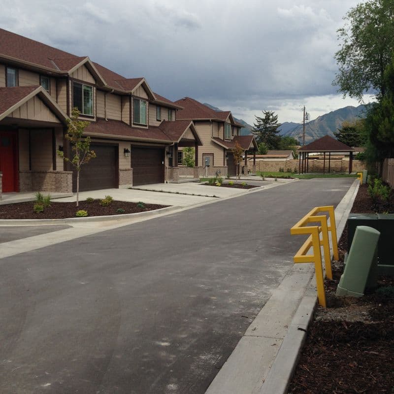 a newly constructed suburban townhome or condo complex with multiple units sharing similar architectural styles. The buildings have brown siding and shingle roofs. In the foreground, there is a concrete street or pedestrian path lined with yellow bollards or barriers. The complex is situated in a mountainous area, with tree-covered hills visible in the background under a partly cloudy sky.