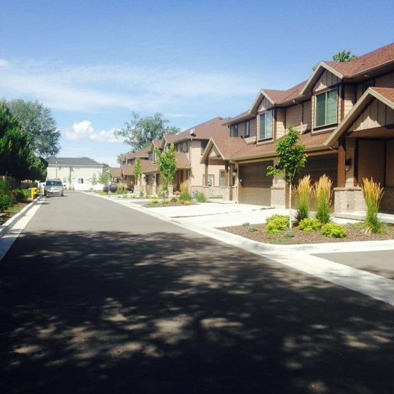 A residential street lined with brown townhouses under a clear blue sky. The street is paved and has concrete curbs with plants and small trees in front of each unit.