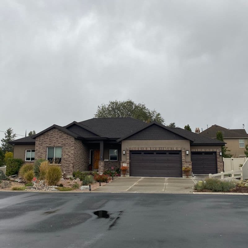 A single-story suburban home with a brick and siding exterior, dark shingle roof, and a two-car garage. The home is landscaped with plants, shrubs, and trees, and there is a concrete driveway leading up to the garage. The photo appears to have been taken on an overcast day.