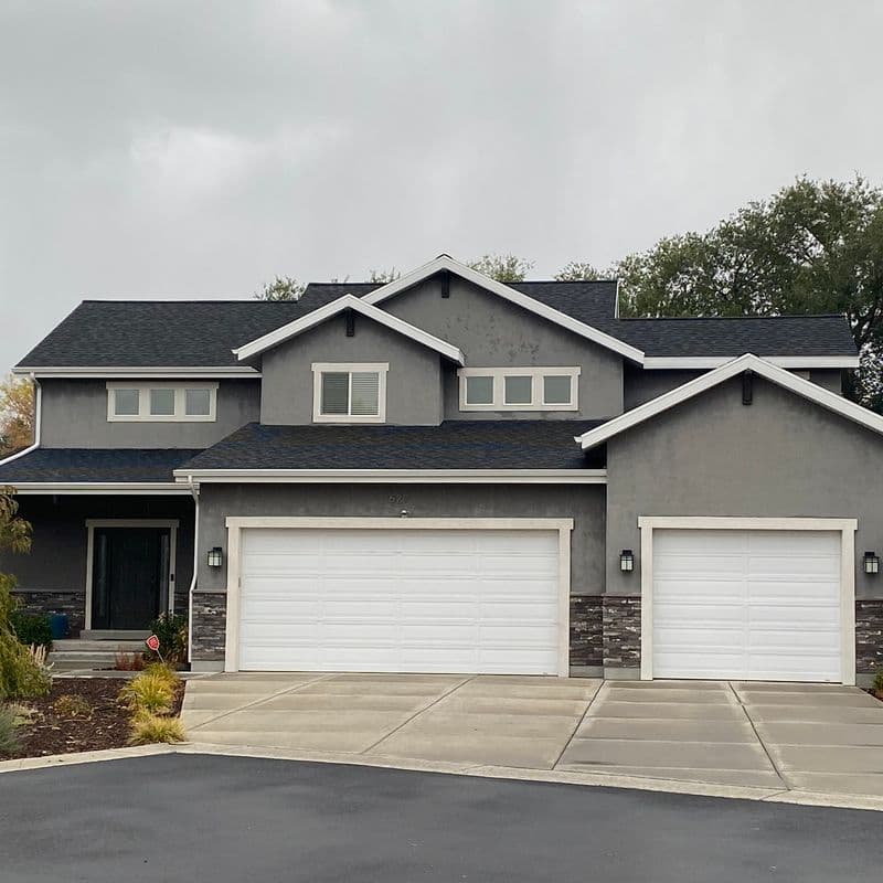 A two-story suburban home with a gray exterior, white trim, and a dark shingle roof. The home has an attached three-car garage with white doors, and there is a concrete driveway leading up to the garage. The landscaping includes trees, shrubs, and plants around the front of the house.