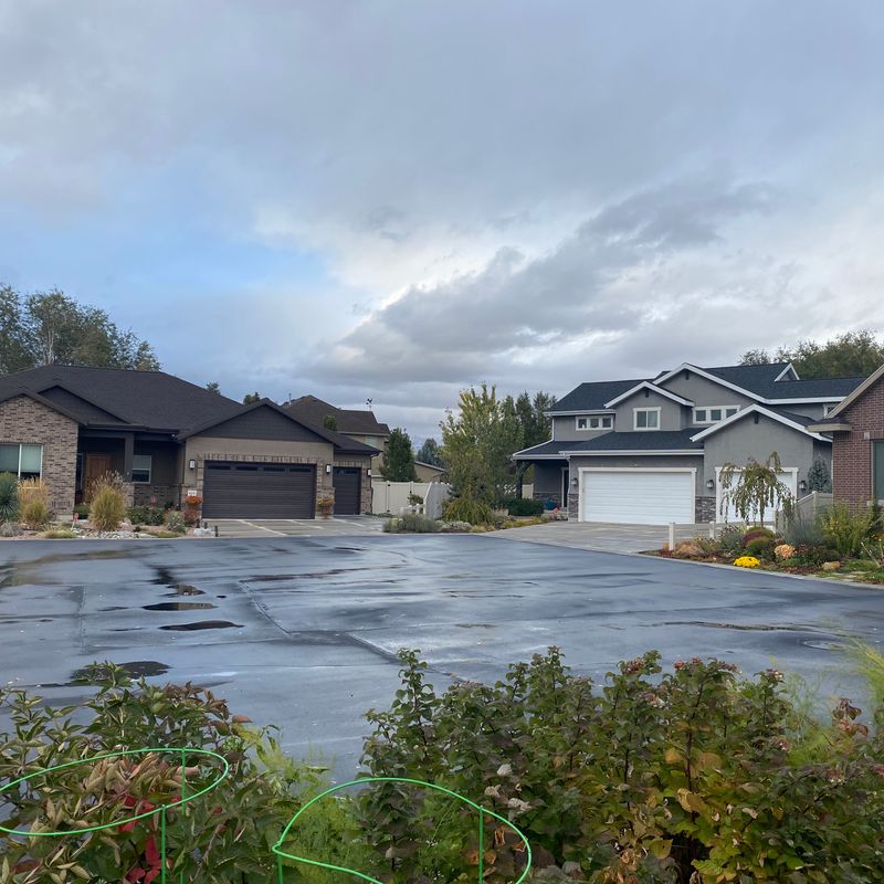 A suburban residential area on a cloudy day. The street is wet, indicating it has recently rained. There are several single-family homes with brick and siding exteriors, garages, and well-maintained front yards with plants and shrubbery. The houses appear to be of similar size and style, suggesting a relatively new housing development or subdivision.