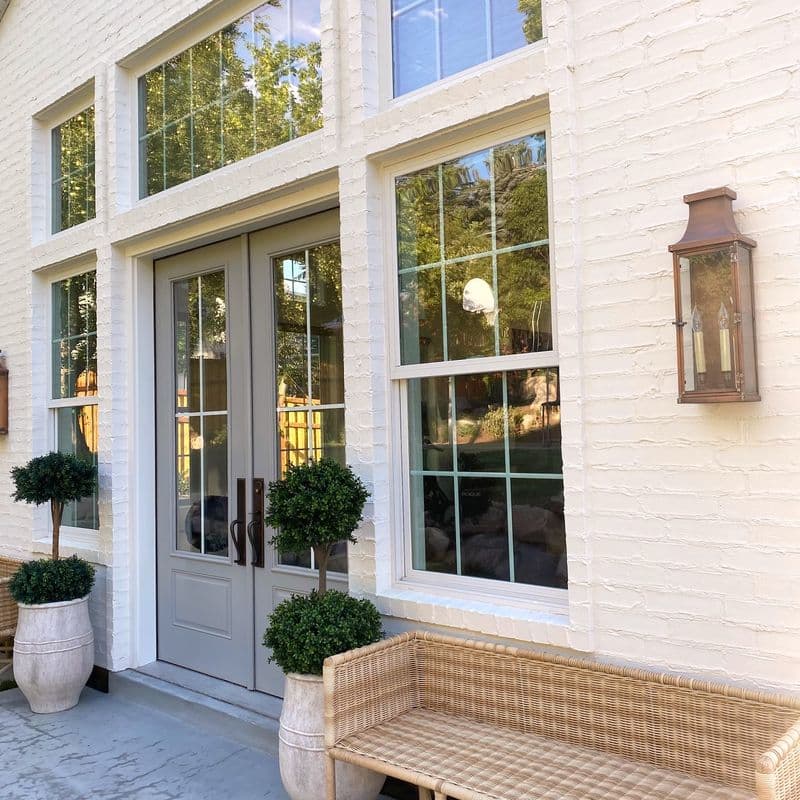 Elegant white brick house exterior with large windows, gray double doors, potted plants, a wicker bench, and a copper wall lantern. The windows reflect lush greenery, creating a bright and inviting entrance.