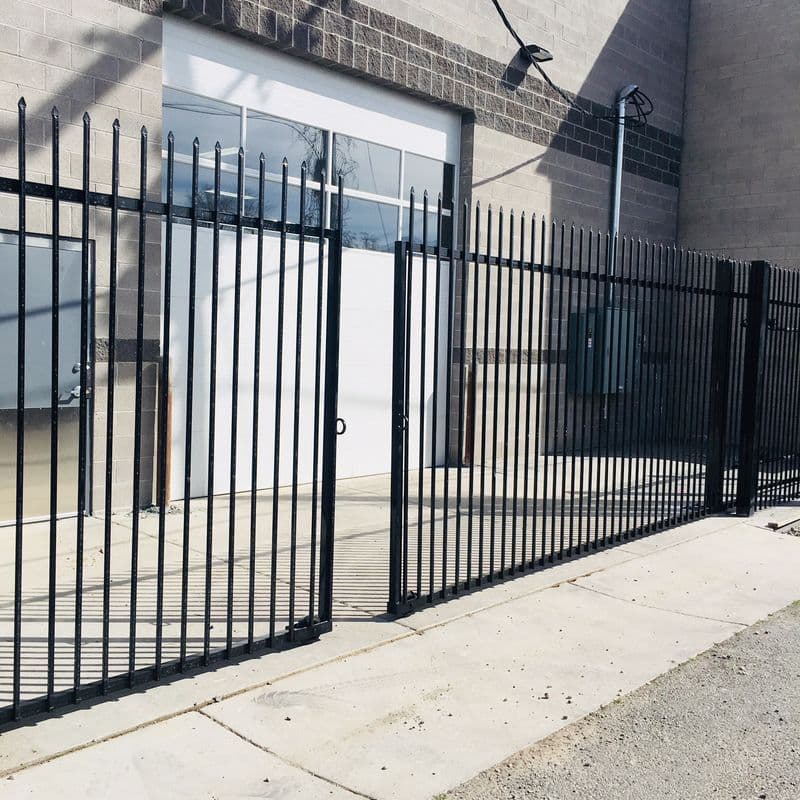 A black metal gate and fence leading to a concrete pathway and garage entrance with a white lifting door. The gate has vertical bars and the fence has a decorative pattern on top.