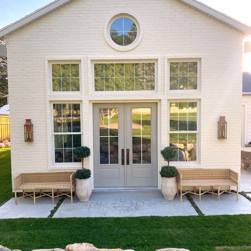 White brick house with large windows, a circular window, and gray French doors. Two wicker benches flank the entrance, with potted topiaries and lantern sconces. A manicured lawn with large rocks borders a concrete patio.